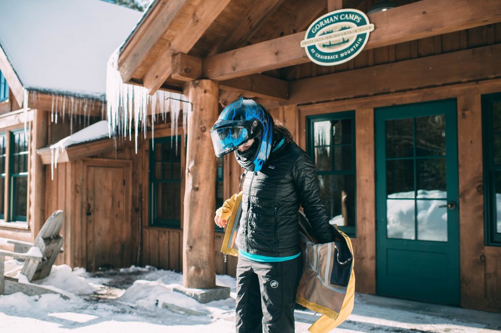 Guest wearting a snowmobile helmet pulls on a jacket in front of Gorman Chairback Cabin.