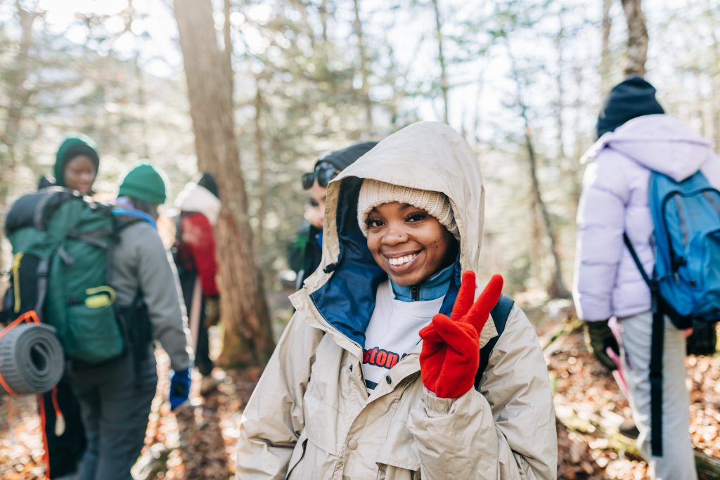 A girl dressed in many layers smiles and gives a peace sign with a red-gloved hand.