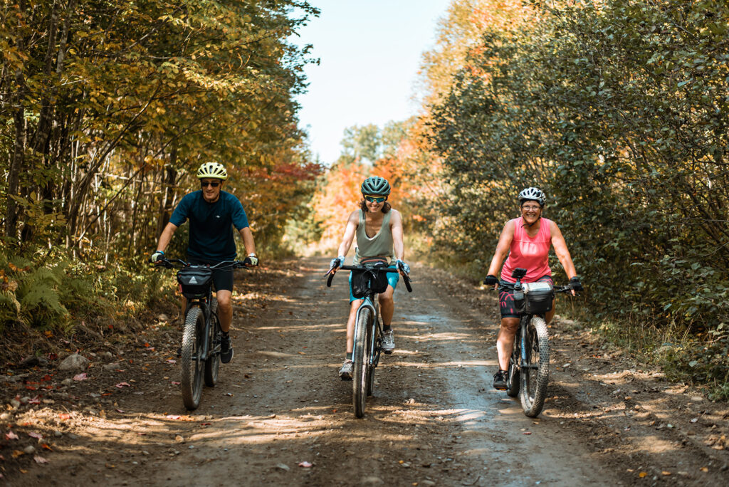 Three smiling people gravel biking on a wide trail in the woods.