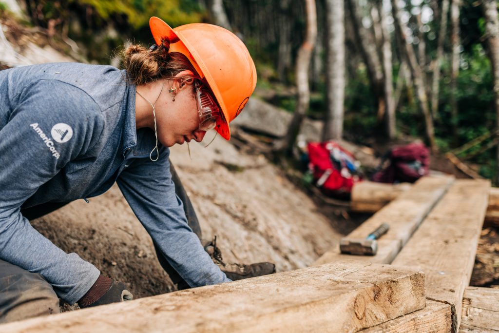 Americorps worker wearing an orange safety helmet on trail with log.