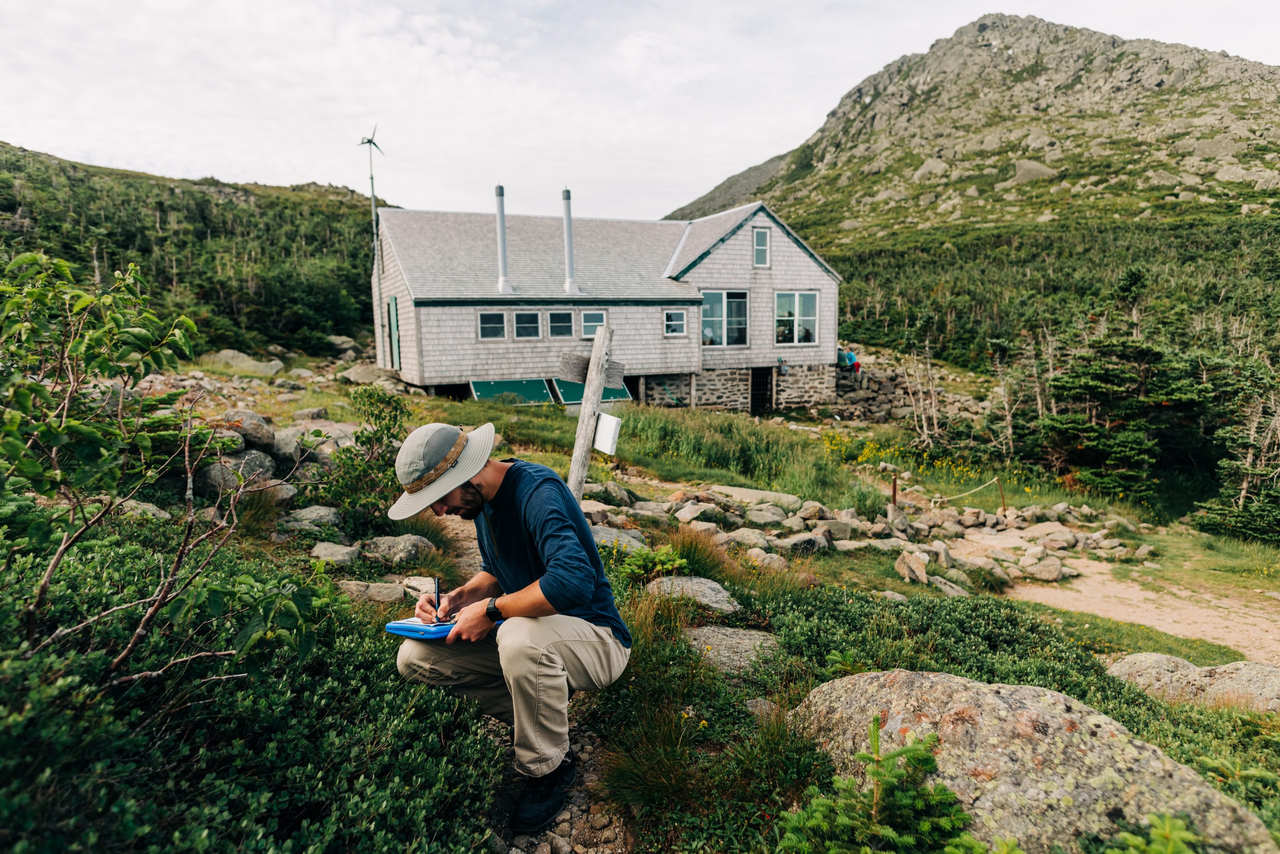 Scientist with a clipboard crouches next to alpine vegetation outside of Madison Spring Hut