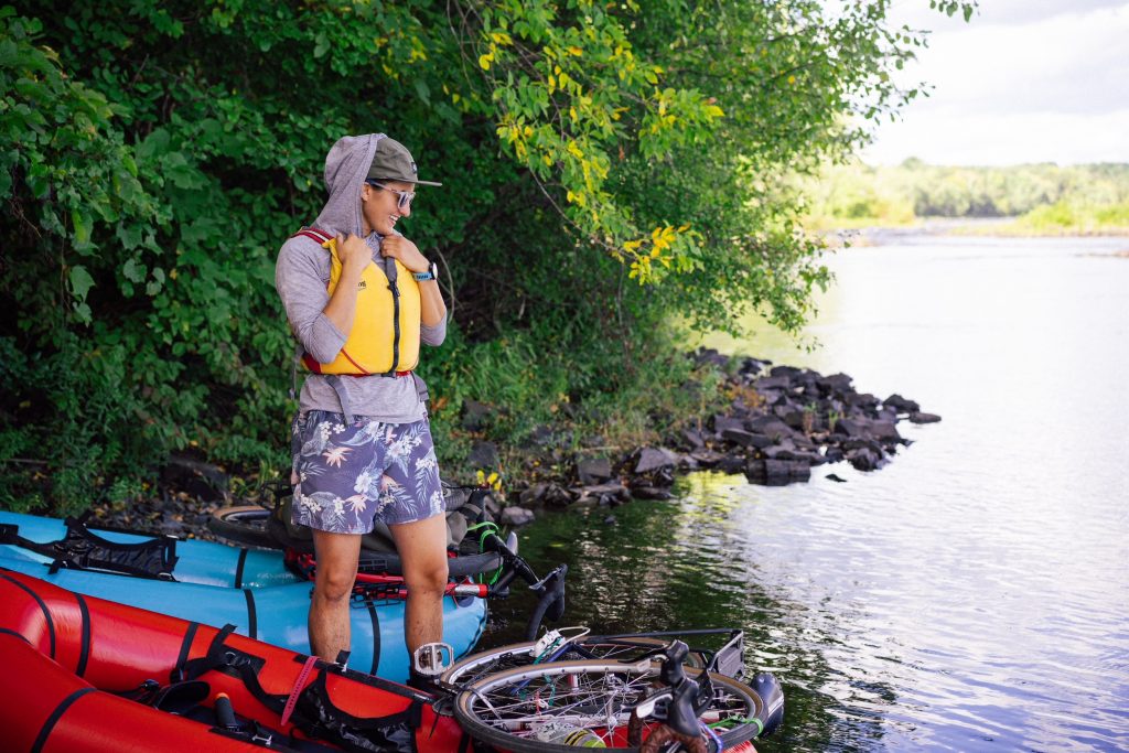 Smiling person standing ankle deep in water next to two rafts loaded with bikes.