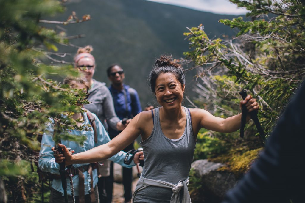 Smiling hiker in gray tank top with trekking poles at the top of a mountain.