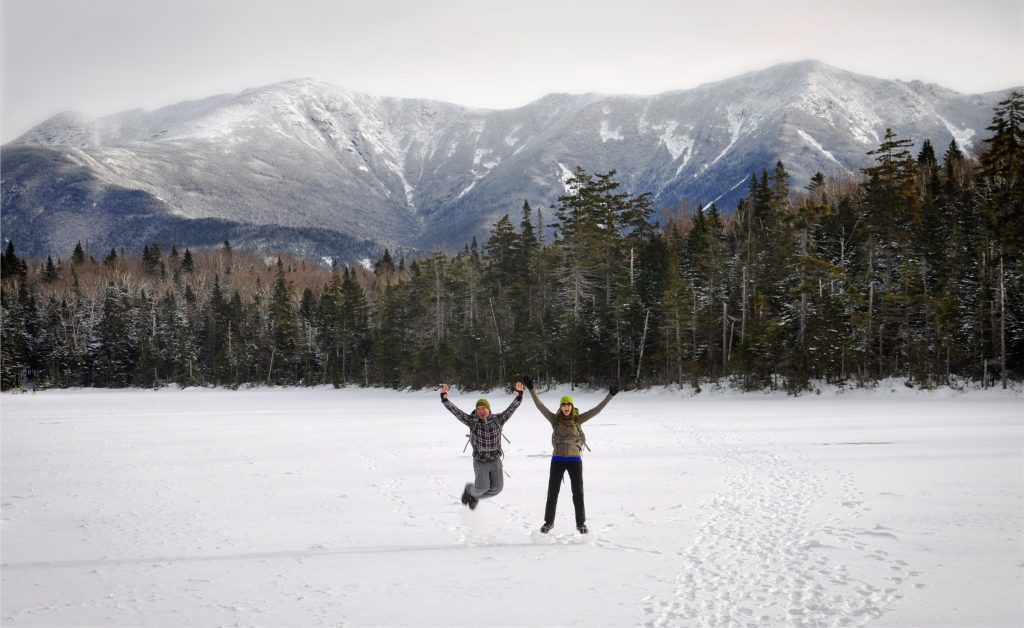 Two people jumping on a frozen lake in front of mountains in winter.