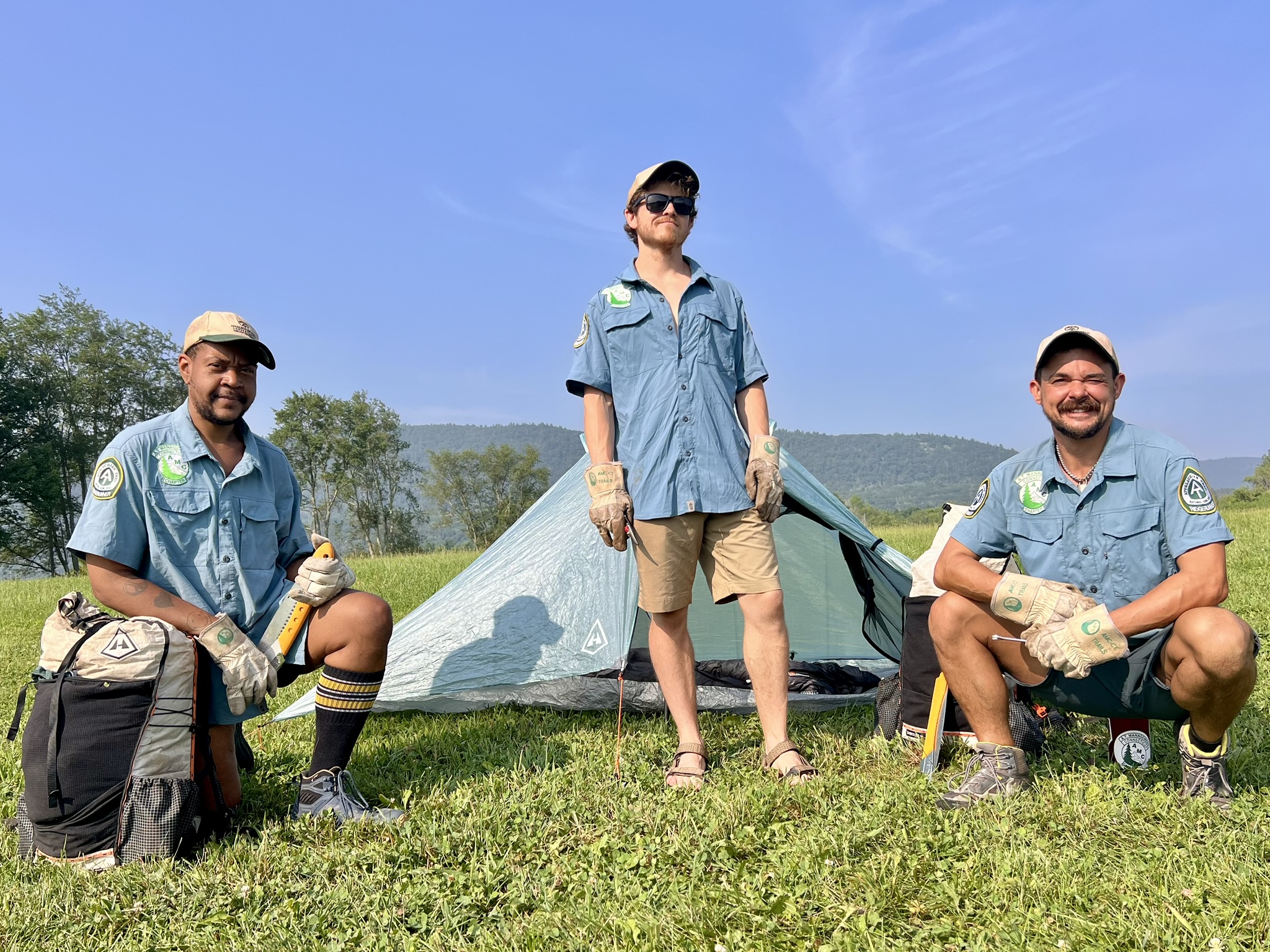 Three ridgerunners sitrting and standing in front of a hyperlite tent on a sunny day.