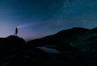 Person with a headlamp standing on a mountaintop and looking at the stars.