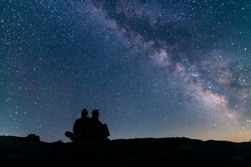 Two people sit back to back and watch the milky way.