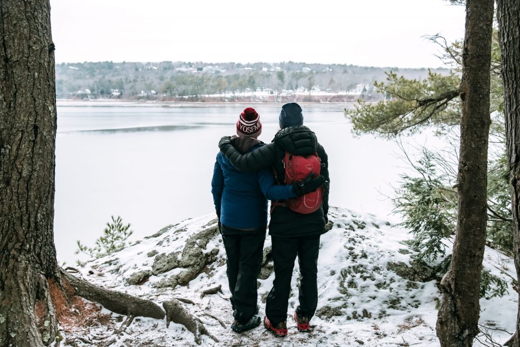 Two people standing in the snow and looking out over water. 