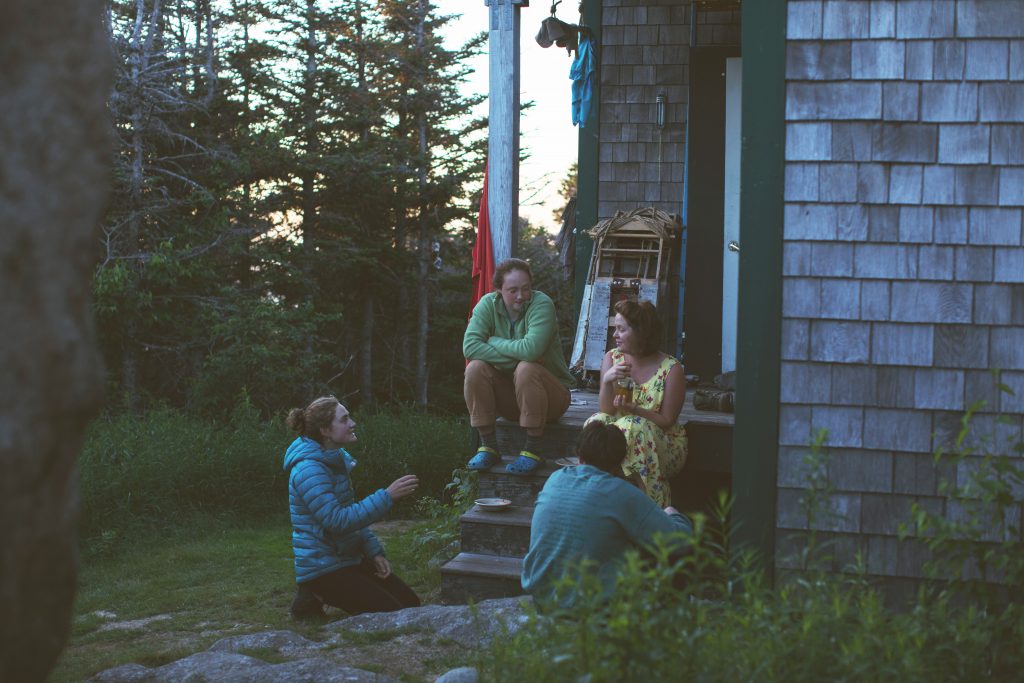 Four people sit on the steps of AMC's Galehead Hut talking. 