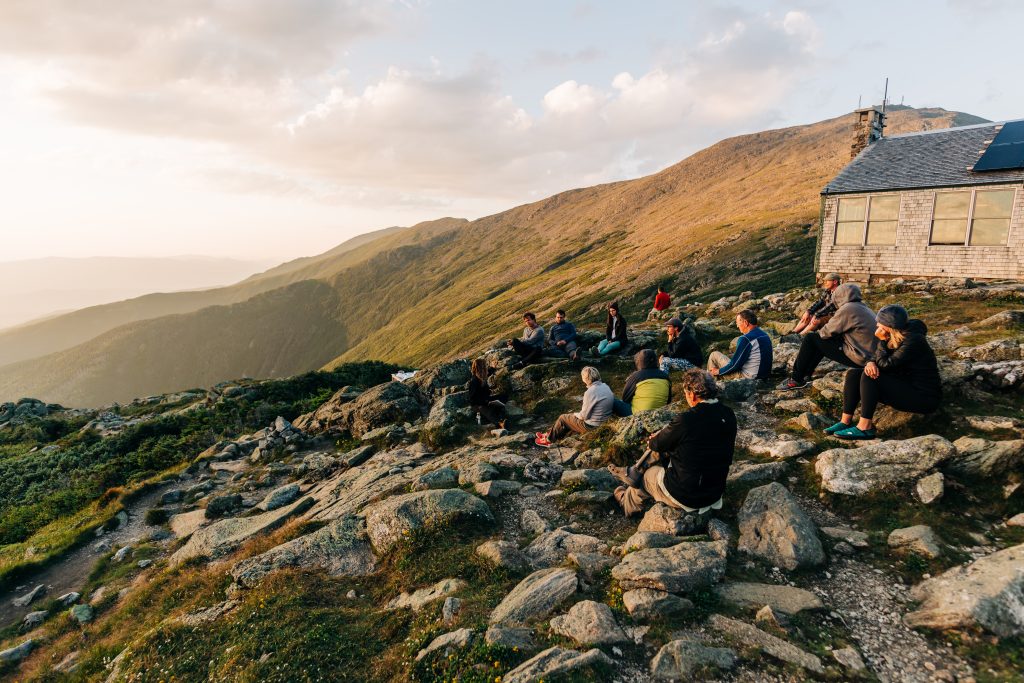 Outside of AMC Lakes of the Clouds Hut, a group of people sit along a rocky edge looking out at a view of the mountains.