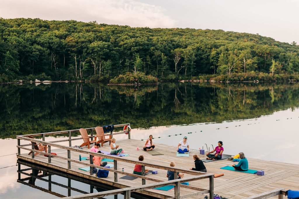 By the lake, a group of people enjoy a yoga class out on a dock at AMC Harriman Outdoor Center.