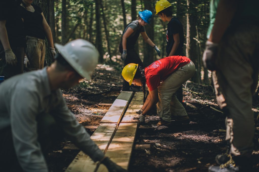 Four volunteers wearing hard hats lay down wooden boards to repair damaged trails on Bay Circuit Trail in Boxford, Massachusetts.
