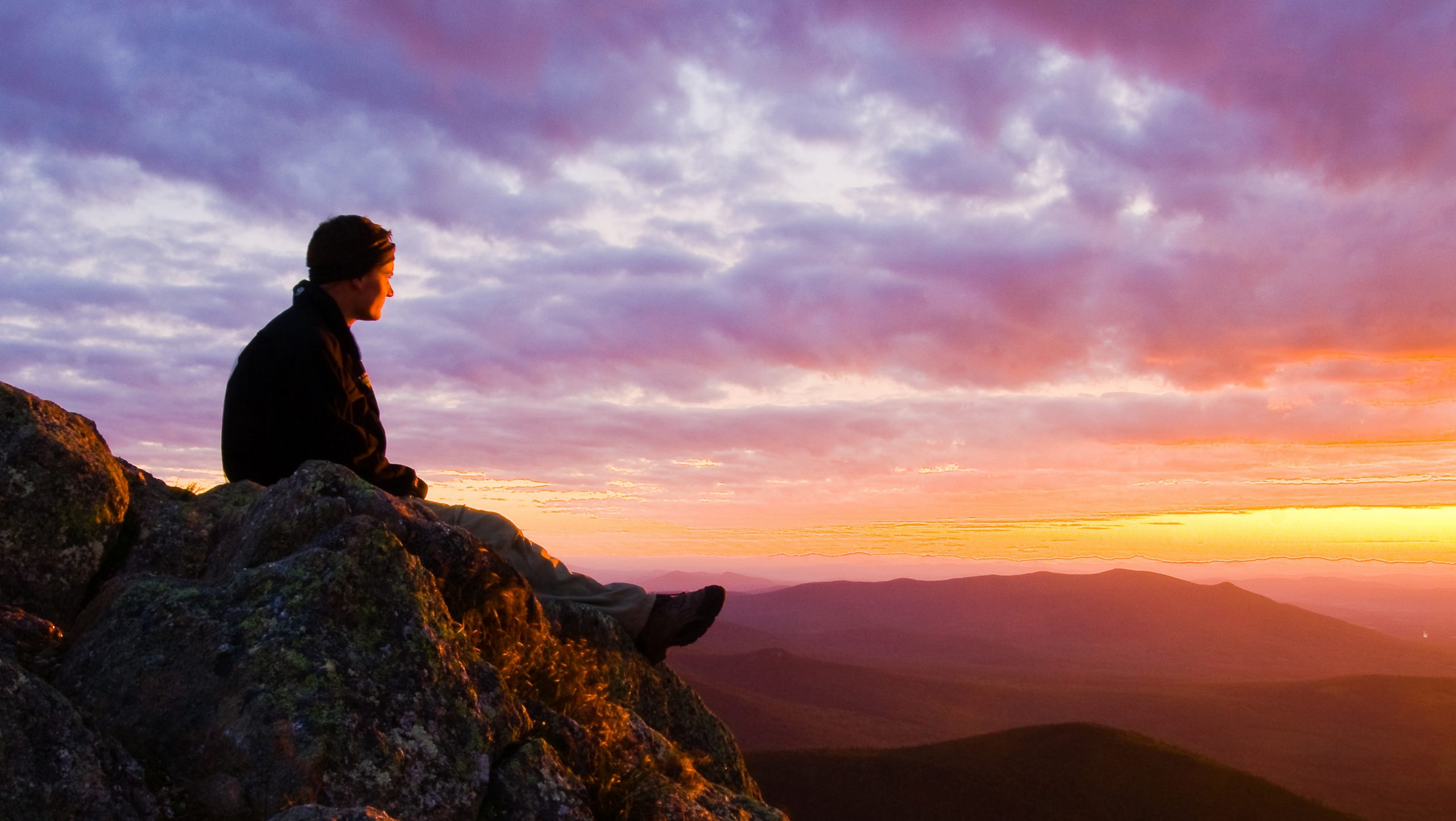 A person sits on a cliff overlooking a view of the sun setting over the White Mountain National Forest.