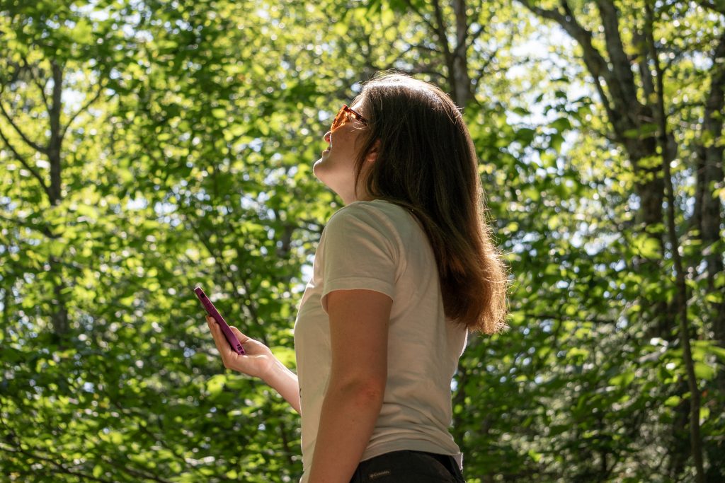 Madelyn Wood in a sunbeam, observing trees and holding an iphone.
