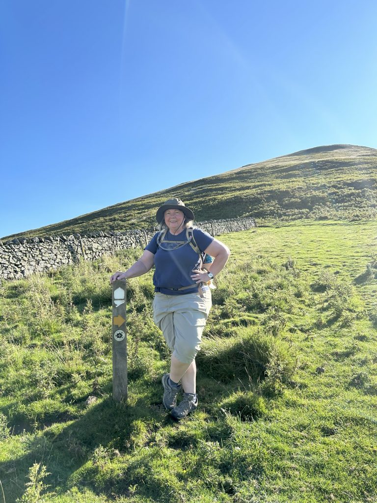 Cathy Poppenwimer stands with one hand on a trail marker on a mountain ridge.