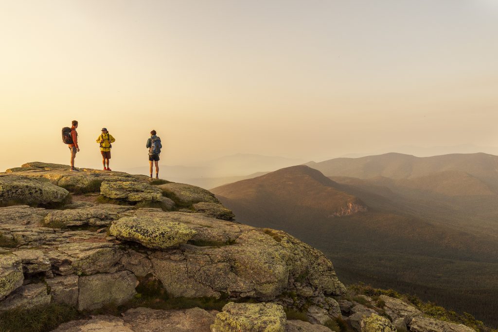 Three people hiking up on Franconia Ridge at dawn, with golden light coming over the mountains.