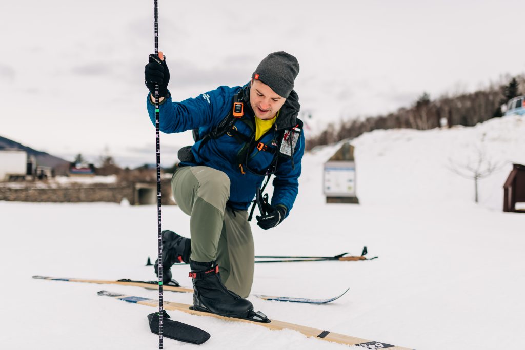 Man in skis kneels with a measuring stick in the snow.