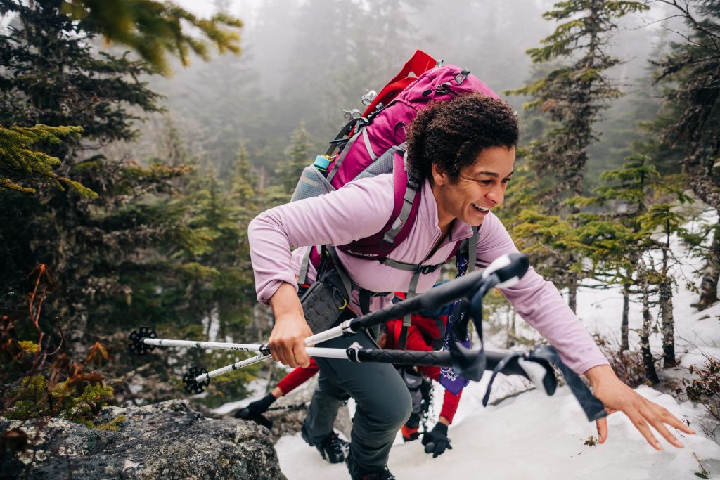 Smiling woman holds poles in her hand while she climbs up a snow rock.