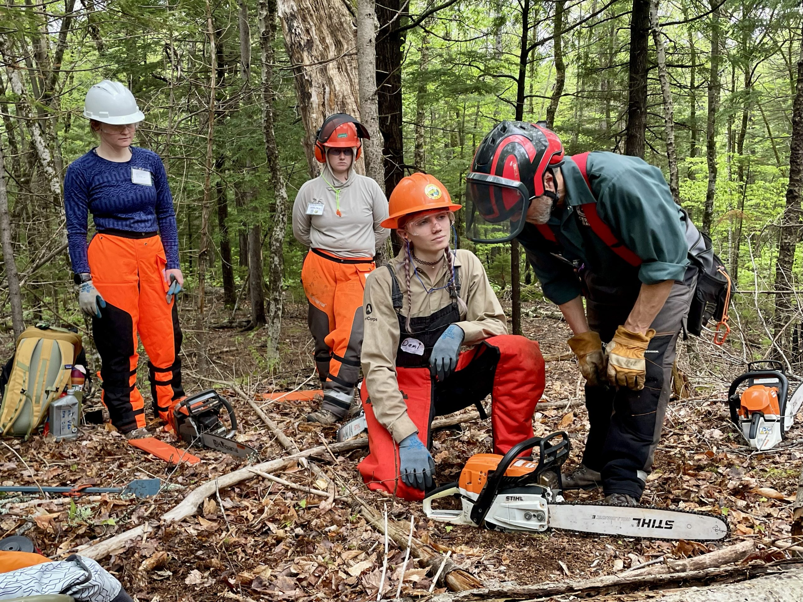 Trail worker with long purple braids kneels by a chainsaw.