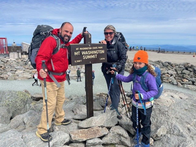A smiling family on top of Mt. Washington with a blue sky behind them.