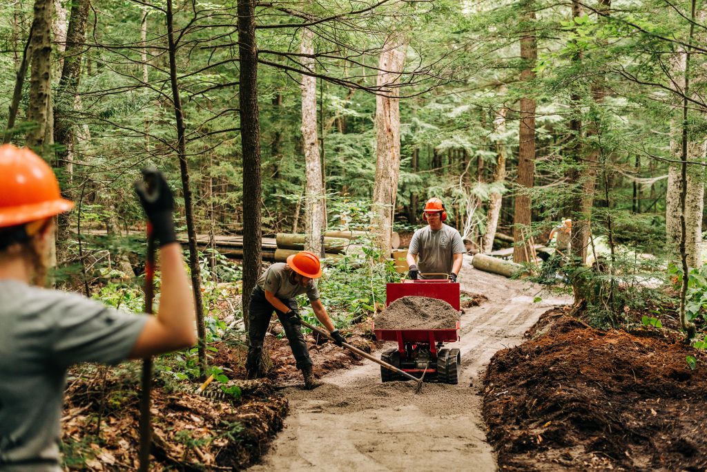 Three adults in orange hard hats use shovels and a wheelbarrow to smooth a portion of the All Persons Trail at AMC Cardigan Lodge.