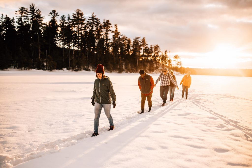 Five people walk through the snow with a bright sun in the background.