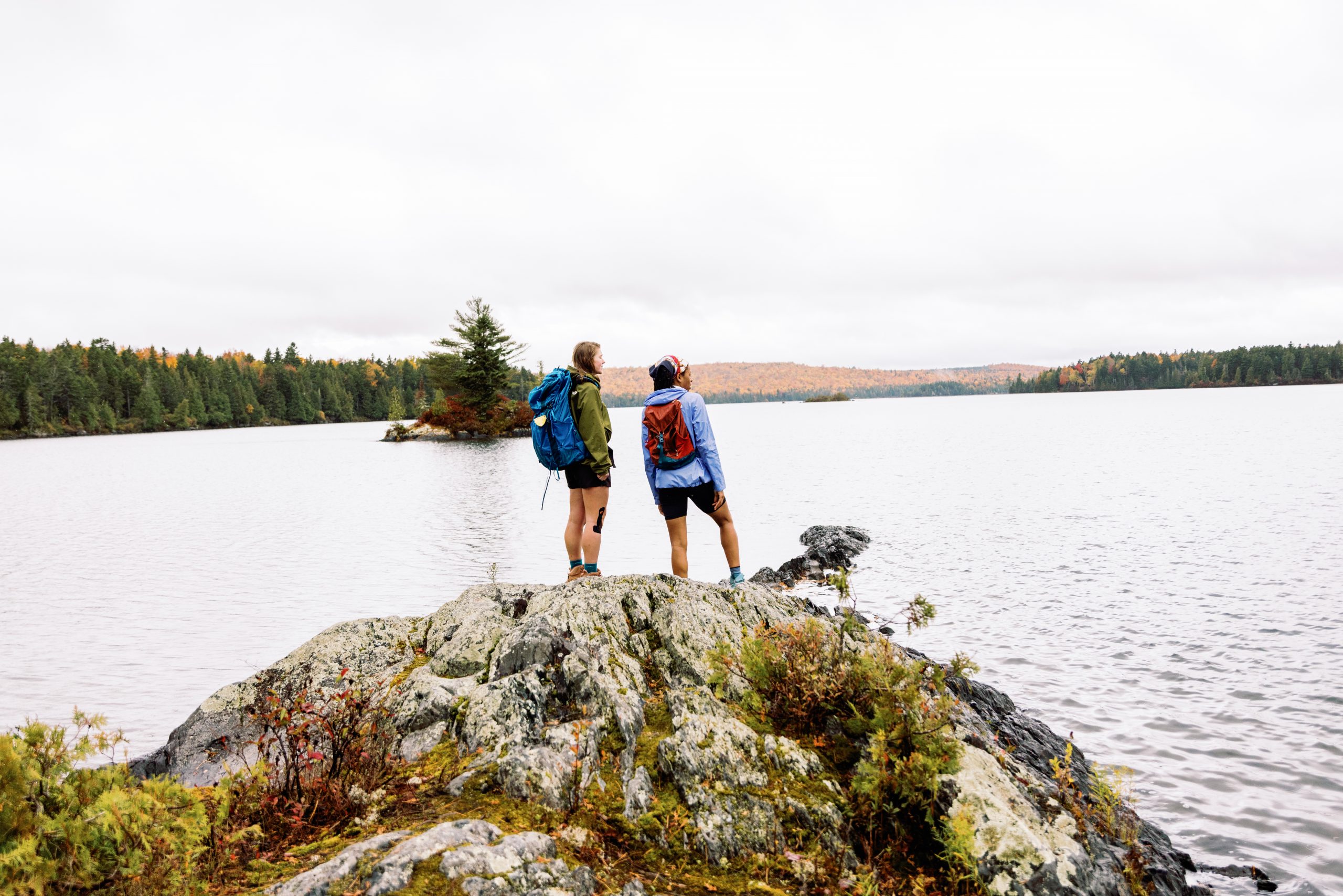 Two people wearing backpacks and standing on a rock by a lake.