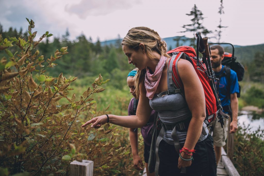 A woman points toward a plant, showing her family what she has discovered, as the group walks along a bridge with water and pine trees in the background.