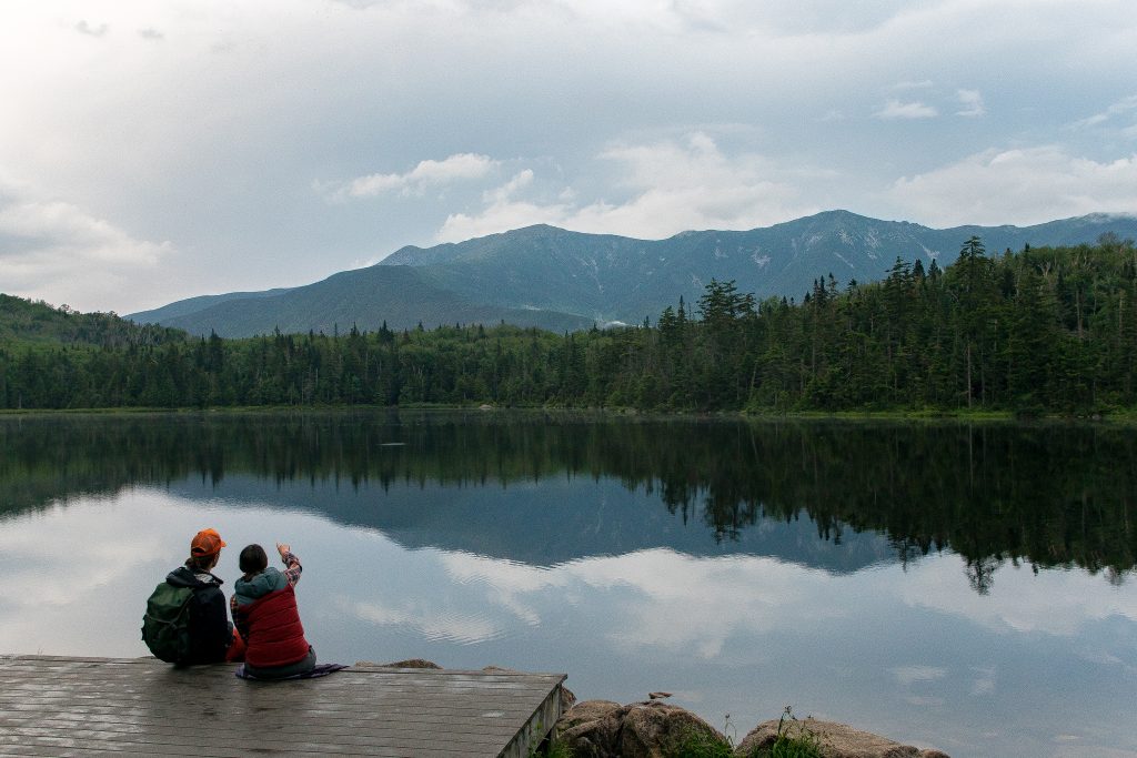 Two people sitting on a dock by a lake and gazing at a mountain.