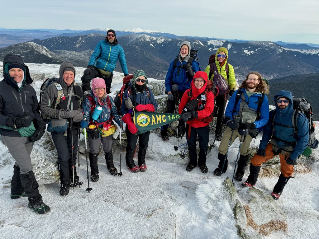 Smiling hikers on top of a snowy mountain holding an AMC 150 flag.