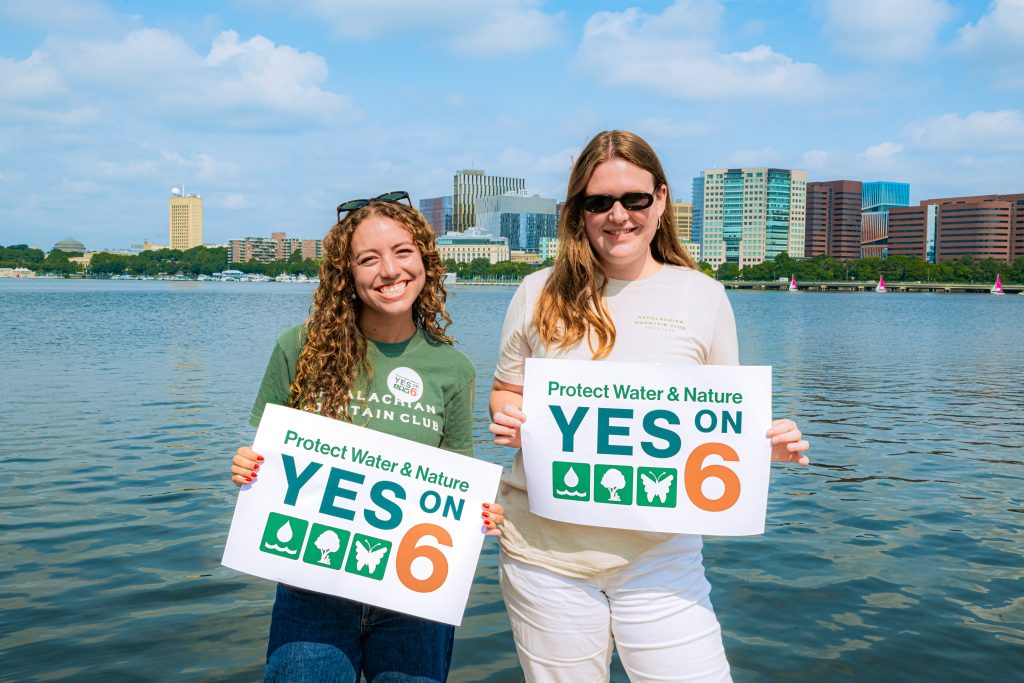 Two people dressed in Appalachian Mountain Club shirts hold signs that read "Protect Water & Nature YES on 6" while standing in front of the Charles River with a view of the city behind them. 