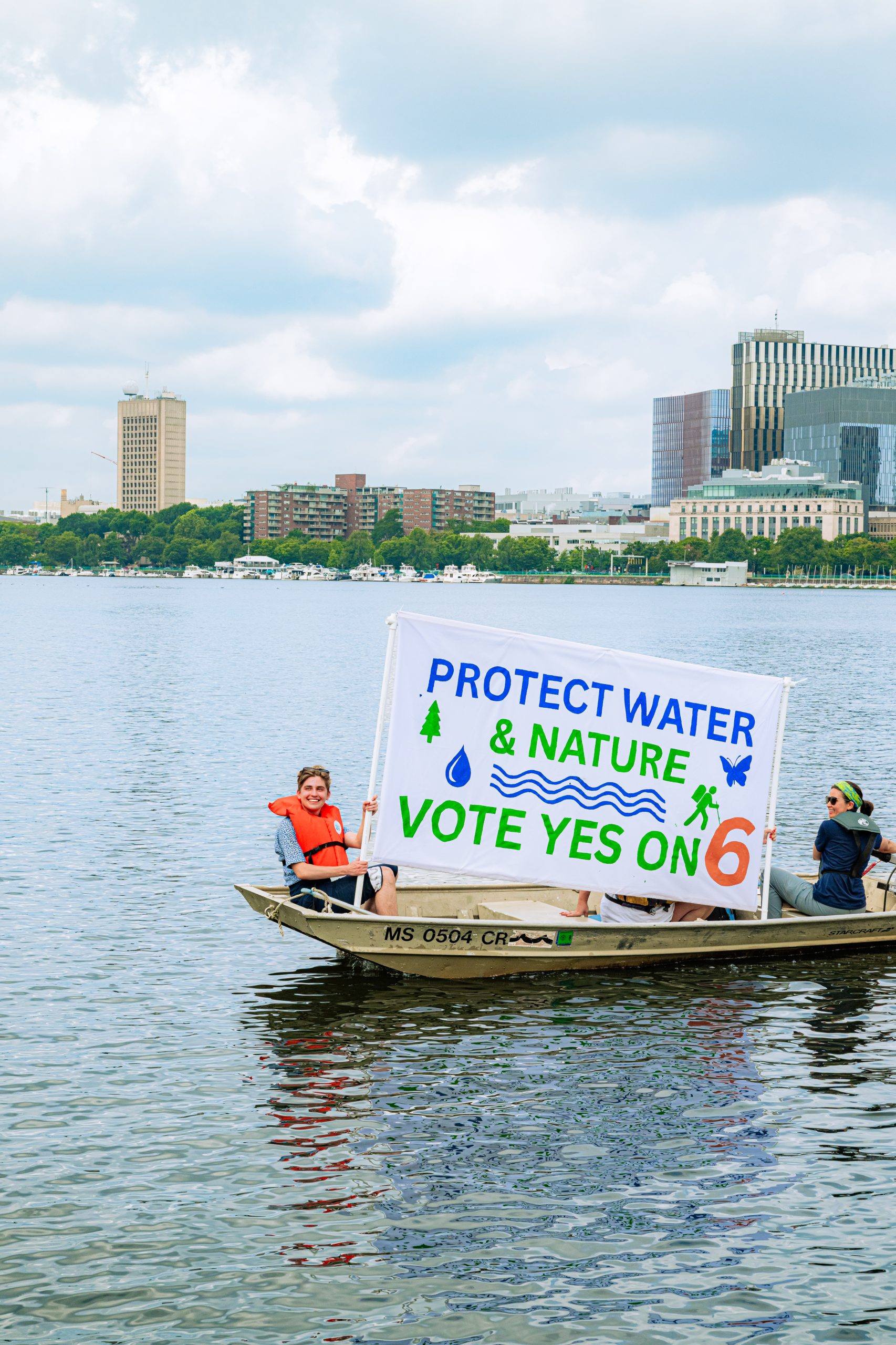 Two people in a boat on the Charles River hold a large YES on 6 sign